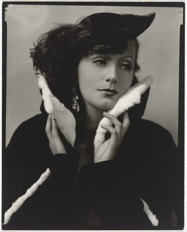A black-and-white portrait of Greta Garbo wearing a dark coat with a white fur collar and a feathered hat, holding the collar up to her chin.