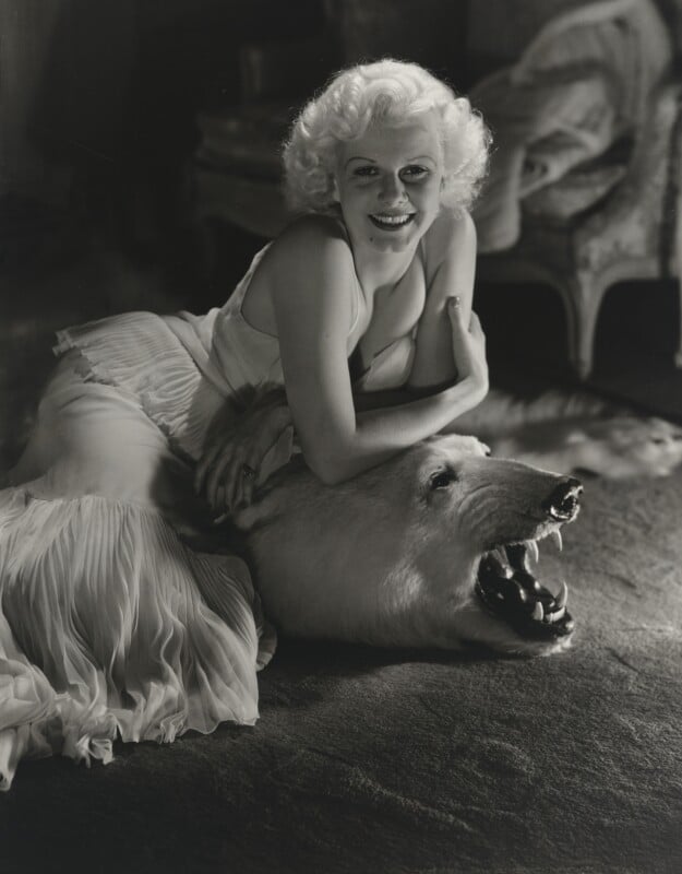 A blonde woman in a light-colored, pleated gown smiles while resting her arms on the head of a taxidermied polar bear rug.