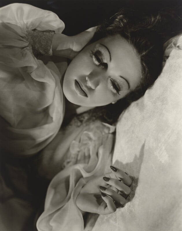 A black-and-white portrait of a woman with long eyelashes lying down, resting her head on her hand.