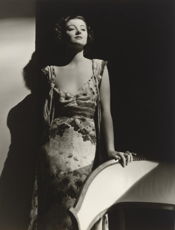 A woman with dark, wavy hair poses in a floral dress, leaning against a chair in a high-contrast, black-and-white portrait.