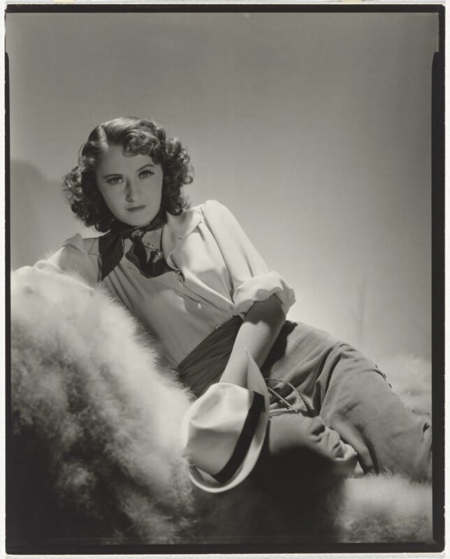 A woman with wavy brown hair, wearing a white collared shirt, a dark neckerchief, and light-colored trousers, reclines on a fur-covered surface while holding a white fedora.