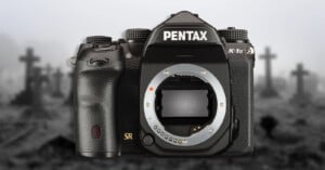 A black Pentax K-1 Mark II DSLR camera body centered against a blurred, grayscale background of a graveyard with crosses.