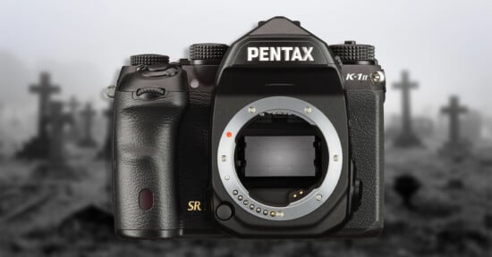 A black Pentax K-1 Mark II DSLR camera body centered against a blurred, grayscale background of a graveyard with crosses.