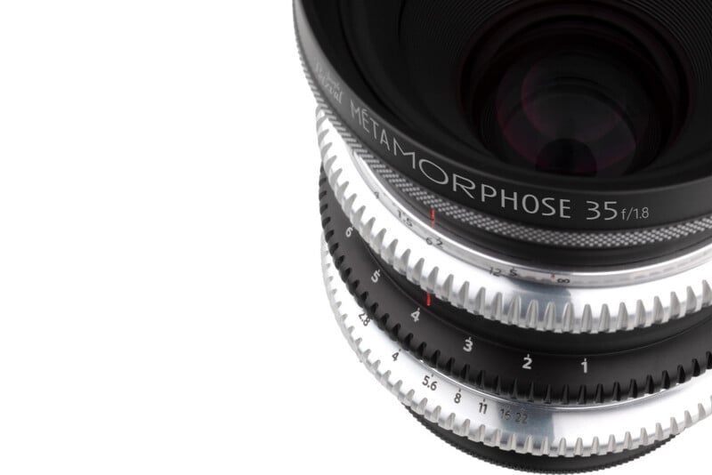 A close-up of a camera lens with silver and black gear-like rings, labeled "METAMORPHOSE 35 f/1.8," against a white background.