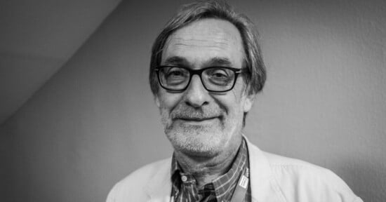 A black and white portrait of a middle-aged man with thinning hair, a beard, and glasses, wearing a collared shirt under a light-colored jacket.