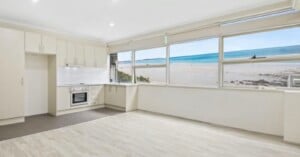 A bright, modern kitchen and living area with light-colored cabinetry, wood-look flooring, and a large window overlooking a beach and ocean.