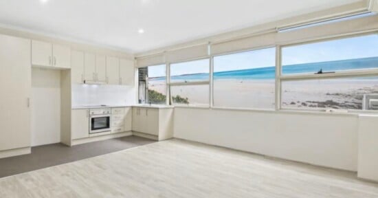 A bright, modern kitchen and living area with light-colored cabinetry, wood-look flooring, and a large window overlooking a beach and ocean.