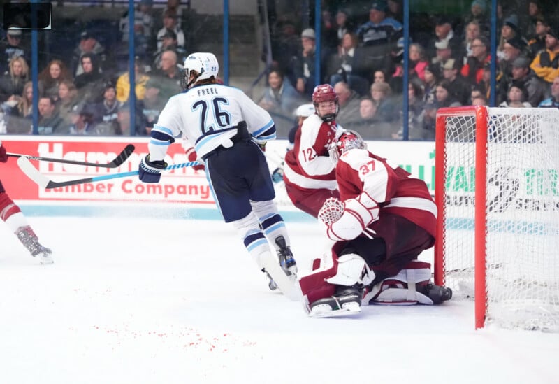 A hockey player in a white jersey with the number 76 skates toward the goal, while the opposing goalie in maroon gear blocks the net.