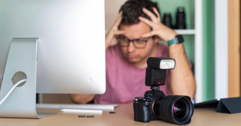 A person wearing glasses sits at a desk with their hands on their head, looking stressed, with a camera and flash in the foreground and a computer monitor to the left.