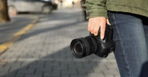 A person wearing a green jacket and blue jeans holds a black DSLR camera in their right hand while standing on a paved sidewalk.