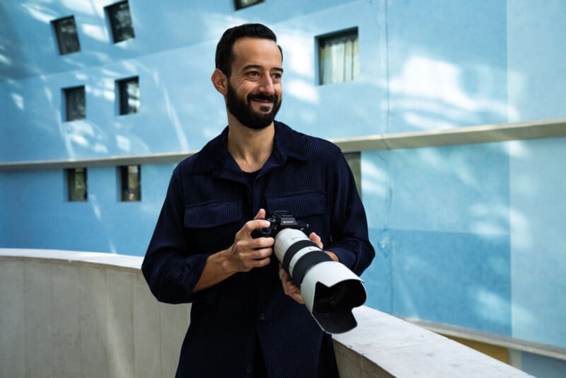 A man with a beard wearing a dark blue button-down shirt holds a camera with a large telephoto lens while standing in front of a blue building with square windows.