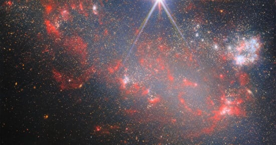 A dense star field featuring glowing red nebulae, clusters of bright stars, and a prominent diffraction spike at the top.