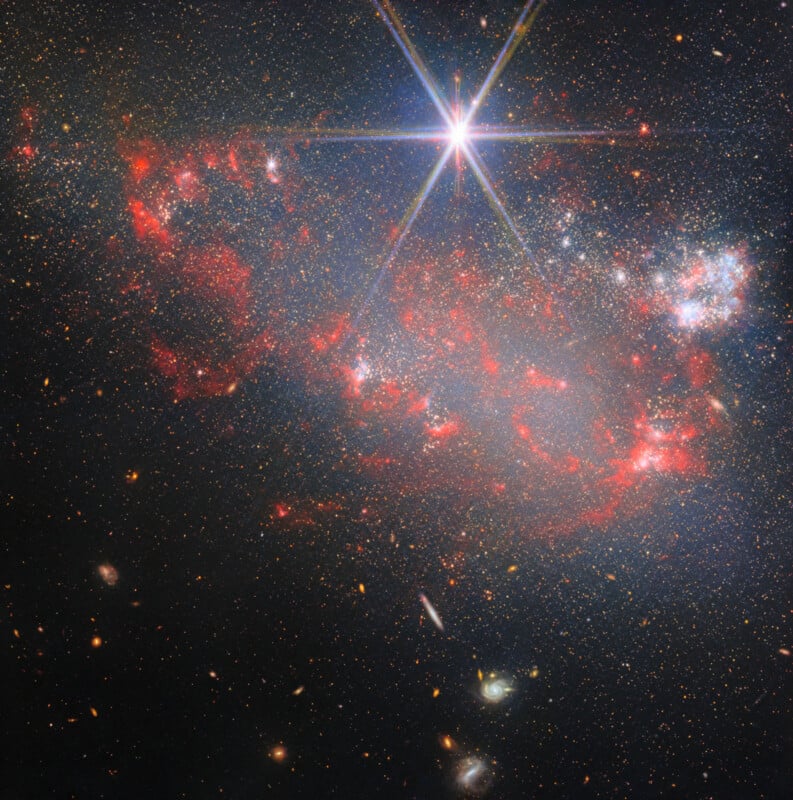A bright star with prominent diffraction spikes shines above a sprawling, irregular galaxy filled with red nebulae and stars.