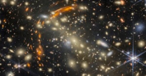 A dense field of galaxies of various shapes and colors, featuring bright stars with diffraction spikes and curved arcs of light.