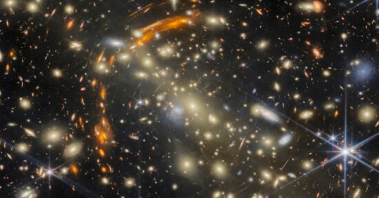 A dense field of galaxies of various shapes and colors, featuring bright stars with diffraction spikes and curved arcs of light.