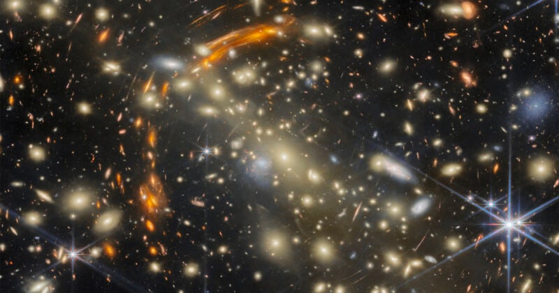 A dense field of galaxies of various shapes and colors, featuring bright stars with diffraction spikes and curved arcs of light.