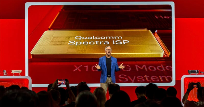A man in a blue blazer stands on a stage in front of a large screen displaying "Qualcomm Spectra ISP" and "X10 5G Modem System" while an audience watches.
