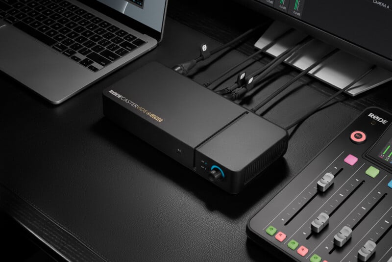 A black RØDECaster Video Core device sits on a desk next to a laptop and a mixing console with several cables connected to the back.