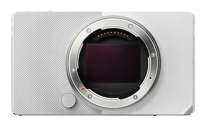 A front-facing silver digital camera body with an exposed sensor and lens mount, featuring a textured grip on the left side.