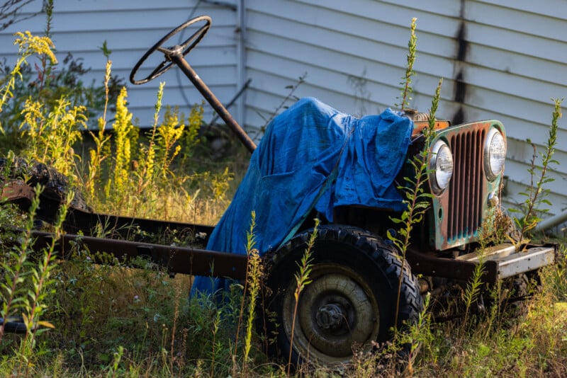 A rusted, partially disassembled vehicle frame with a Jeep-style front grille and headlights sits in a field of tall weeds, covered by a blue tarp.