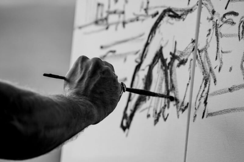 A close-up, black and white shot shows a person's hand holding a thin brush and sketching dark, abstract lines onto a white canvas.