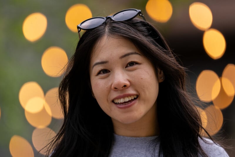 A smiling woman with long dark hair and sunglasses resting on her head poses against a background of blurred golden lights.