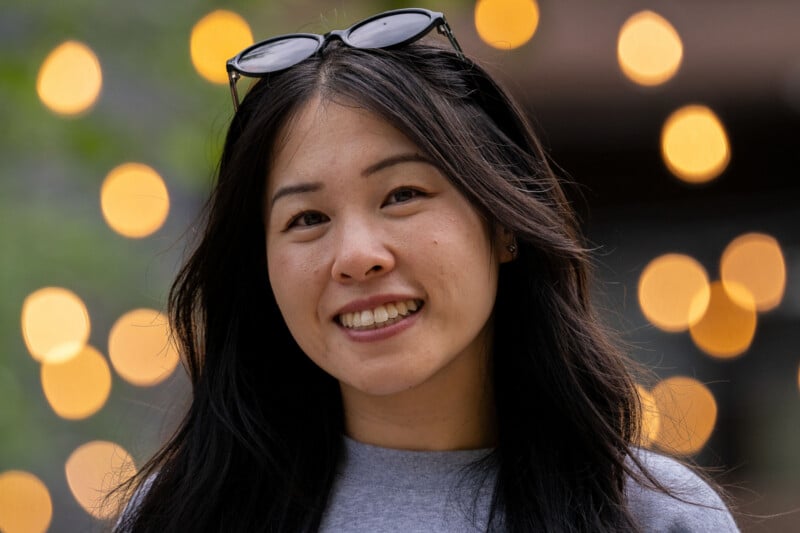 A smiling woman with long dark hair and sunglasses perched on her head stands in front of a background of blurred golden lights.
