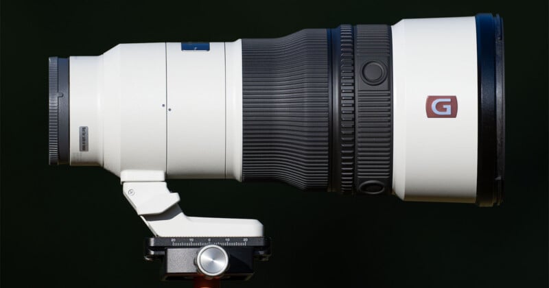 A white telephoto camera lens with a black zoom ring and a "G" logo is mounted on a tripod head against a dark background.