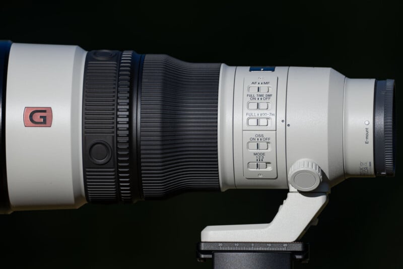 A side view of a white telephoto camera lens mounted on a tripod, featuring various focus and stabilization switches on its side.
