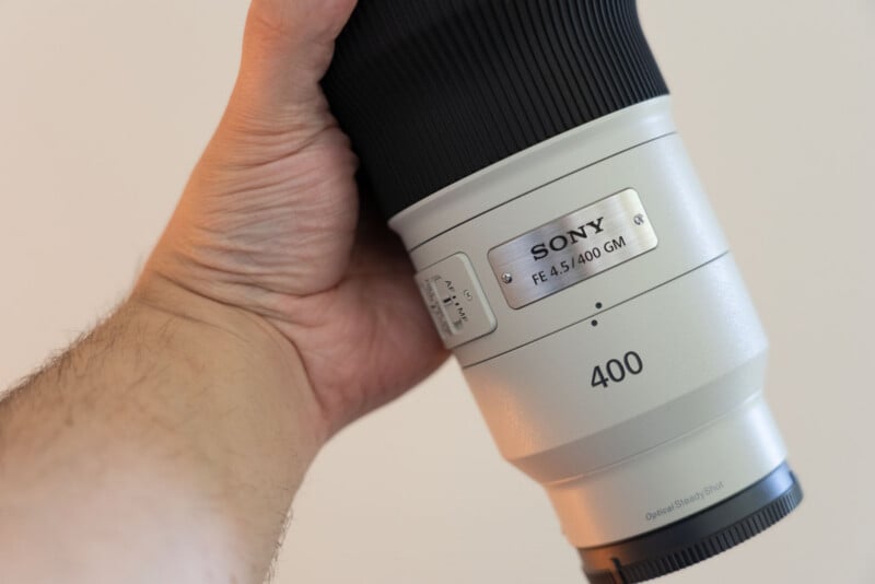 A person's hand holds a white Sony FE 4.5/400 GM lens against a plain background.