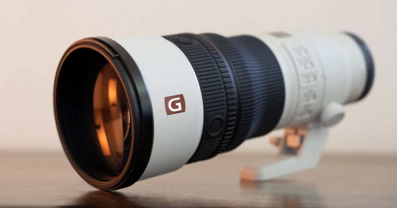 A white telephoto camera lens with a black zoom ring and a prominent "G" logo sits on a wooden surface against a plain background.