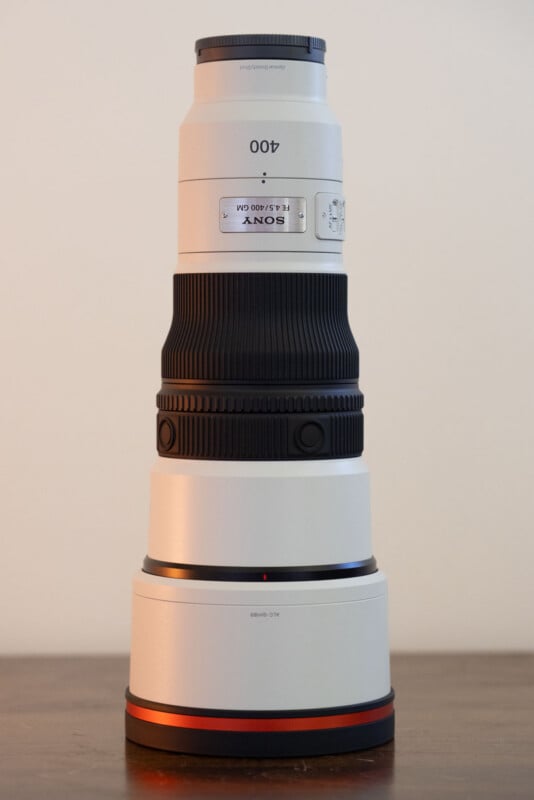 A white Sony FE 4.5/400 GM telephoto lens stands vertically on a wooden surface against a plain background.
