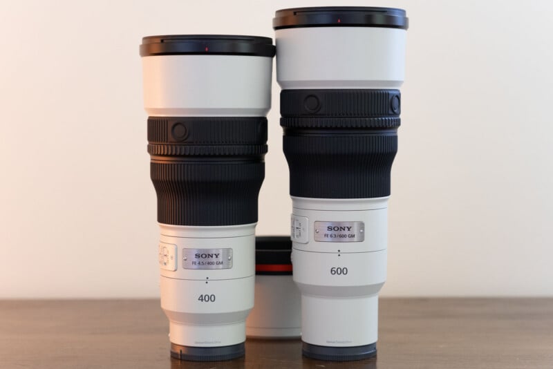 Two white Sony telephoto lenses stand upright on a wooden surface, with a 400mm lens on the left and a 600mm lens on the right.