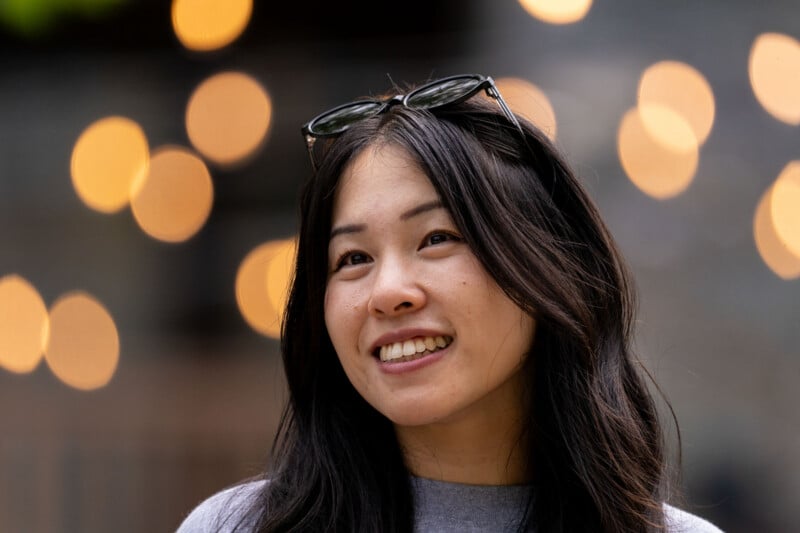A young woman with long dark hair and sunglasses perched on her head smiles while looking off to the side against a background of blurred golden lights.