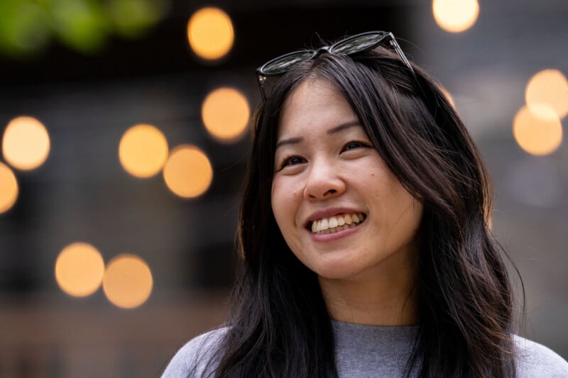 A smiling young woman with long dark hair and glasses perched on her head stands in front of blurred, glowing bokeh lights.