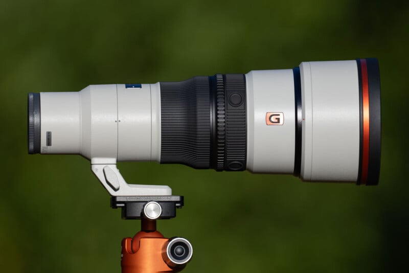 A long, white telephoto camera lens with a black focus ring and a prominent orange "G" logo, mounted on an orange tripod head against a blurred green background.