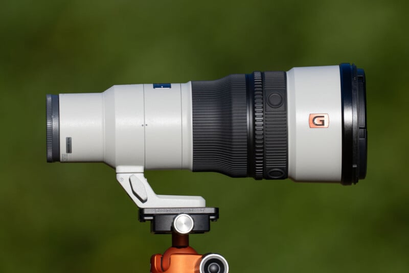 A white telephoto camera lens mounted on a tripod head against a blurred green background.