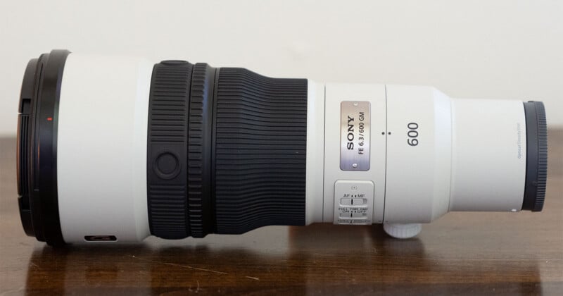 A white Sony FE 6.3/600 GM telephoto camera lens sits horizontally on a dark wooden surface against a plain white background.