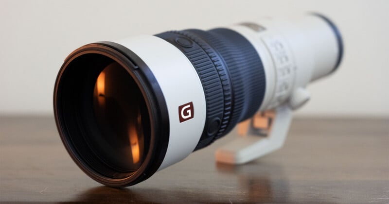 A white telephoto camera lens with a black zoom ring and a red square logo featuring a white letter G, resting on a wooden surface.