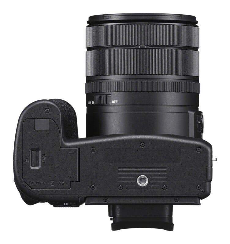 Bottom view of a black digital camera with a lens attached, showing the battery compartment door, tripod mount, and aperture click switch.