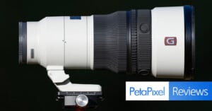 A white telephoto camera lens mounted on a tripod head against a black background, with a PetaPixel Reviews logo in the corner.
