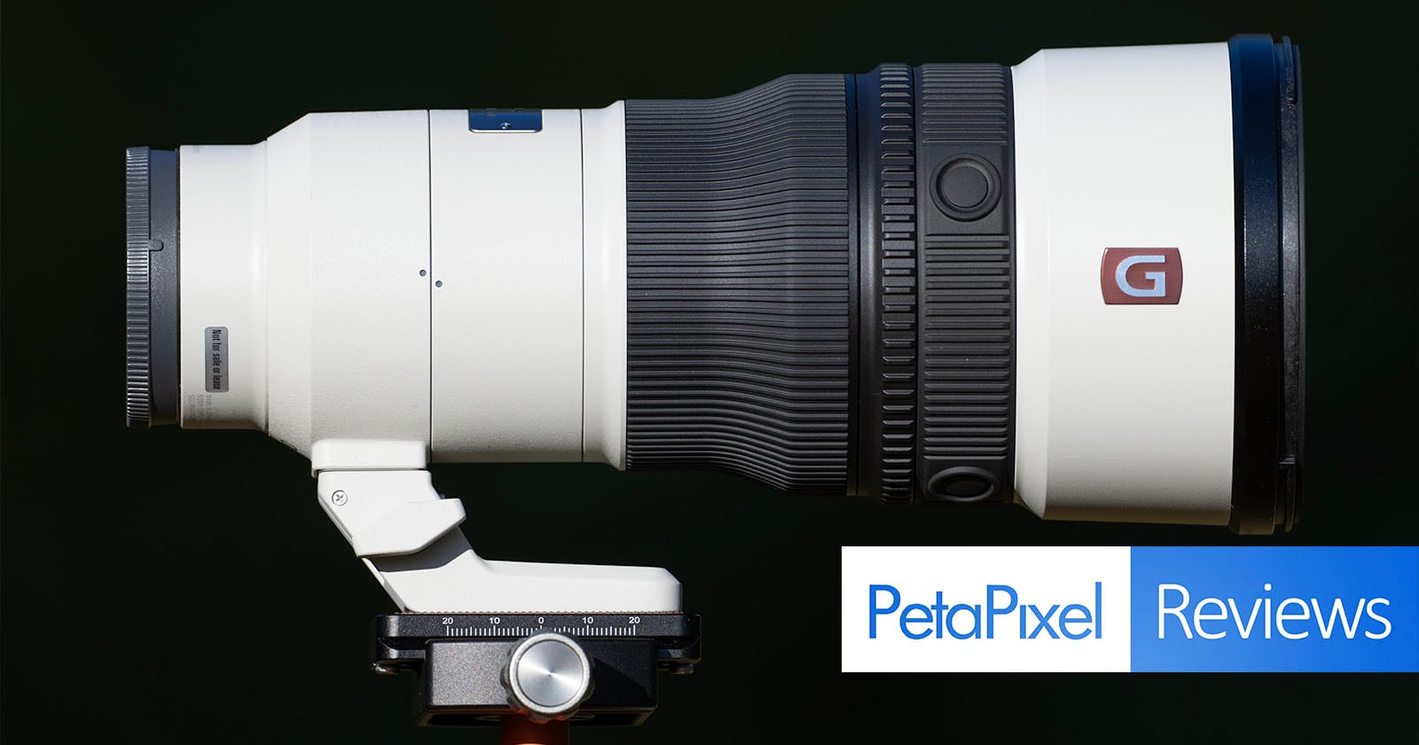 A white telephoto camera lens mounted on a tripod head against a black background, with a PetaPixel Reviews logo in the corner.