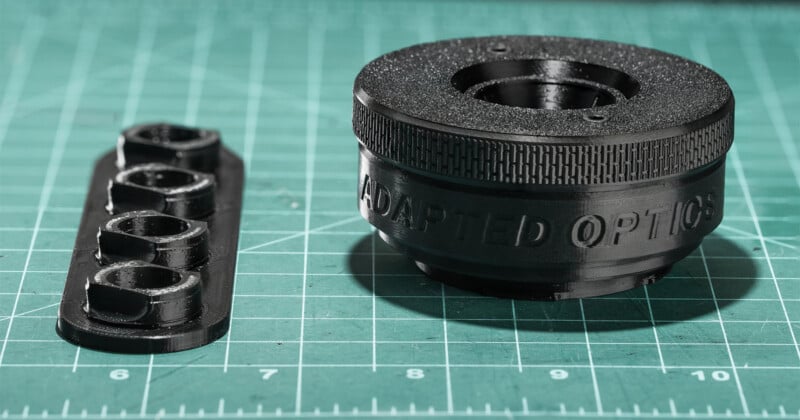 A 3D-printed black circular lens adapter labeled "ADAPTED OPTICS" sits next to a small rectangular strip containing four integrated nut-shaped fasteners on a green cutting mat.