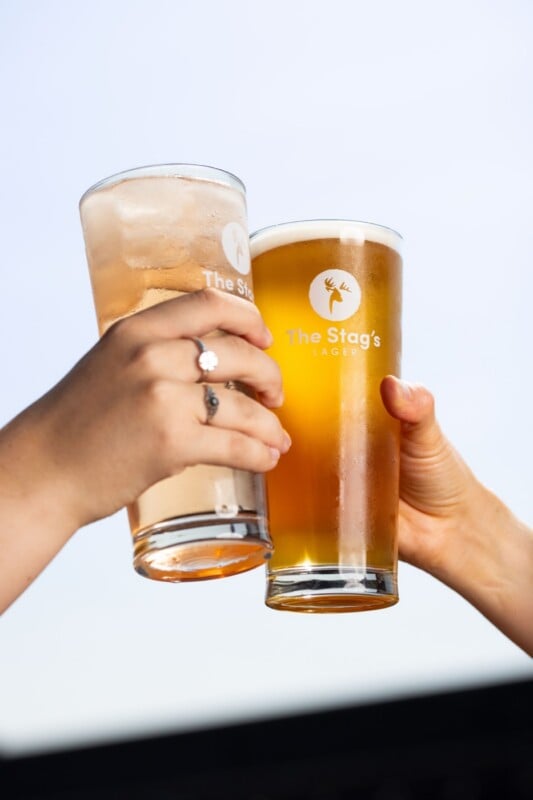 Two hands hold up glasses of beer and a mixed drink to toast against a bright, light-colored background.