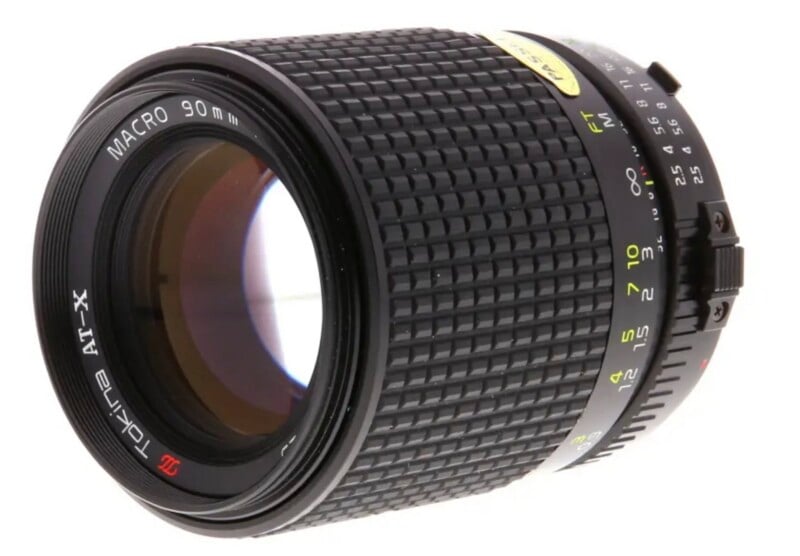 A black Tokina AT-X 90mm macro camera lens shown at an angle against a white background.