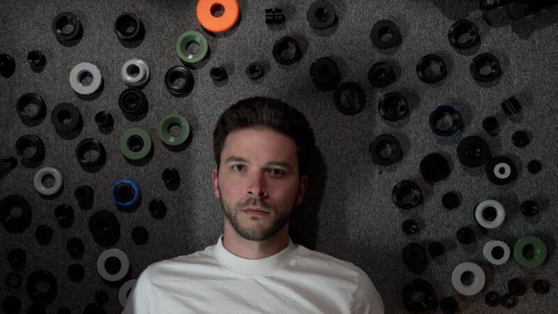 A man with a beard and a white shirt lies on a gray carpet surrounded by numerous circular plastic rings and components.