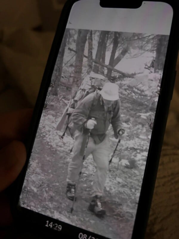 A person wearing a bucket hat and a large backpack hikes through a wooded area while using trekking poles, as seen on a smartphone screen.