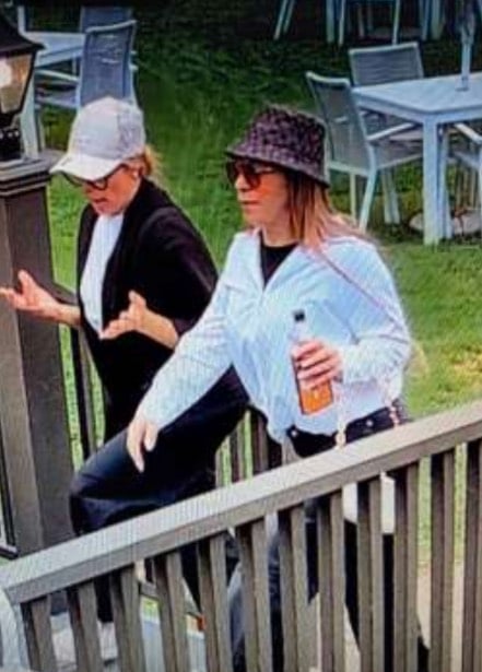 Two women walk up wooden stairs outdoors, one wearing a light-colored baseball cap and black jacket, the other wearing a patterned bucket hat and white button-down shirt while holding a bottle.
