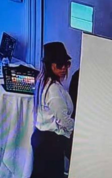 A person with long hair wears a dark bucket hat, sunglasses, and a white long-sleeved shirt, standing near a table with a laptop.