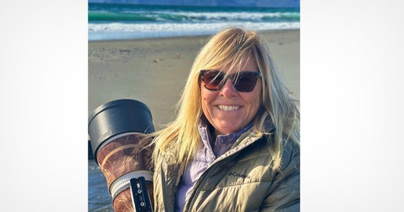 A smiling woman with blonde hair and sunglasses stands on a beach holding a large camera lens with a camouflage cover.
